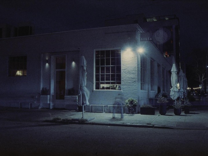 A blurred figure walks past a building with large windows and covered outdoor umbrellas at night.
