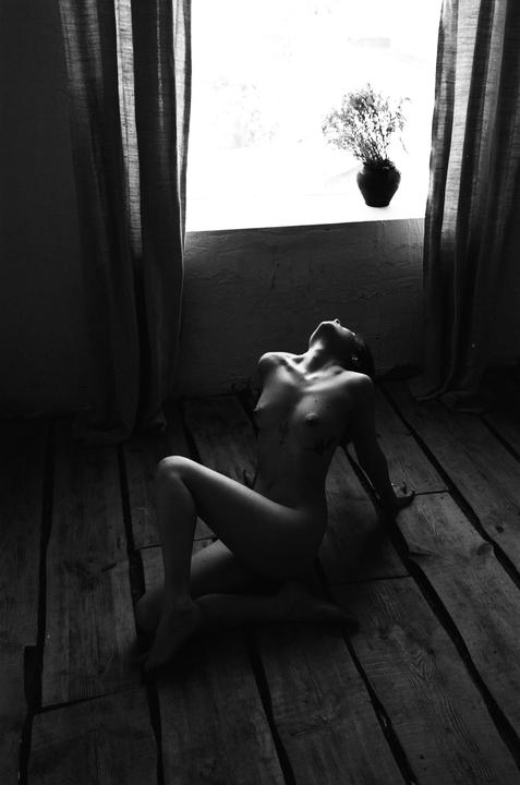 A nude woman sits on a wooden floor with her head tilted back towards a bright window.