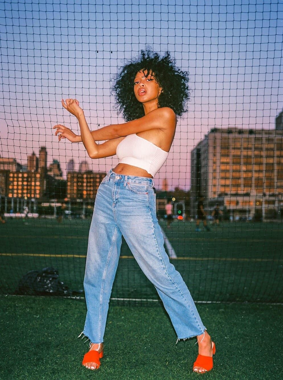 A woman with curly hair poses in front of a net on a grassy field with buildings in the background.