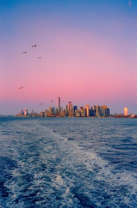A cityscape with tall buildings is visible across a body of water with a boat wake, under a sky transitioning from pink to blue.