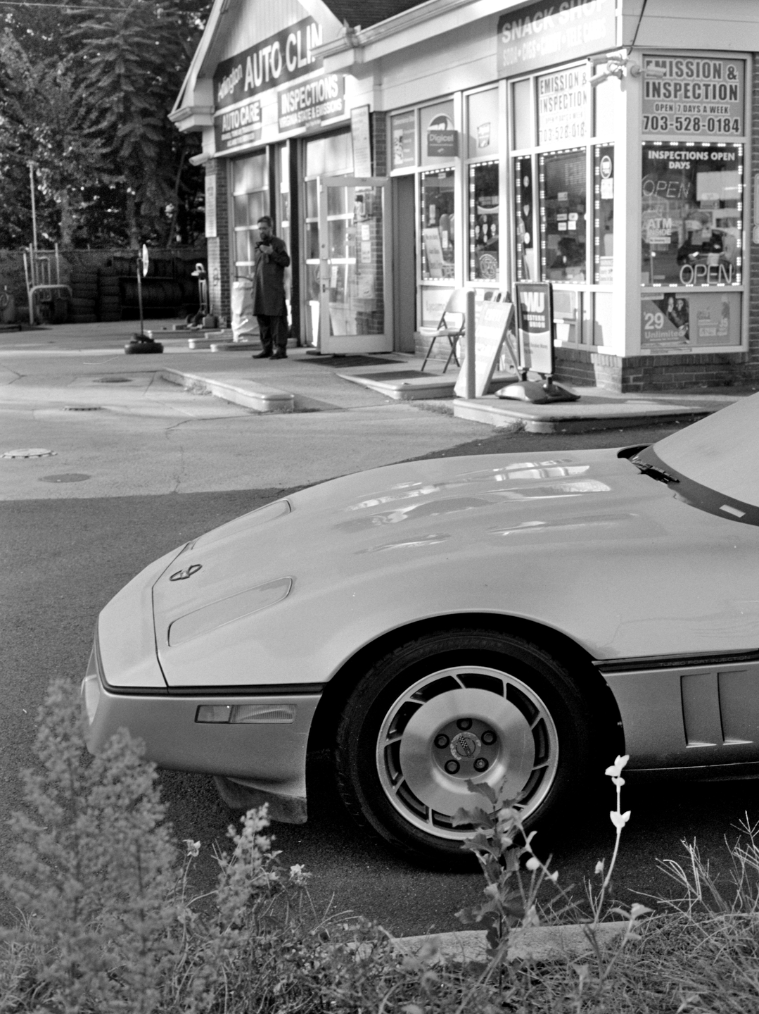 A man stands outside an auto repair shop with a car parked in the foreground.
