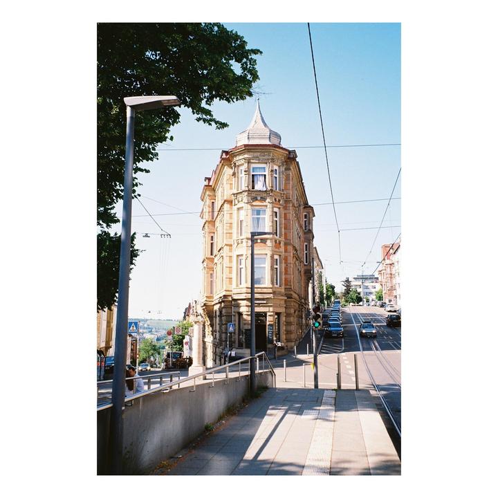 A tall, ornate building with a pointed roof stands at a street corner with cars and tram tracks visible on a sunny day.