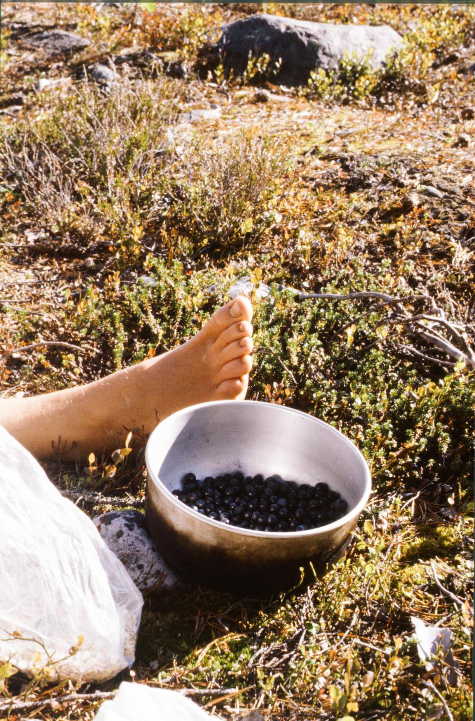 A person's foot rests on the ground next to a bowl filled with blueberries.