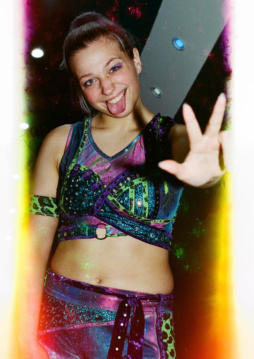 A woman in a colorful costume with glittery makeup sticks out her tongue and holds up her hand.