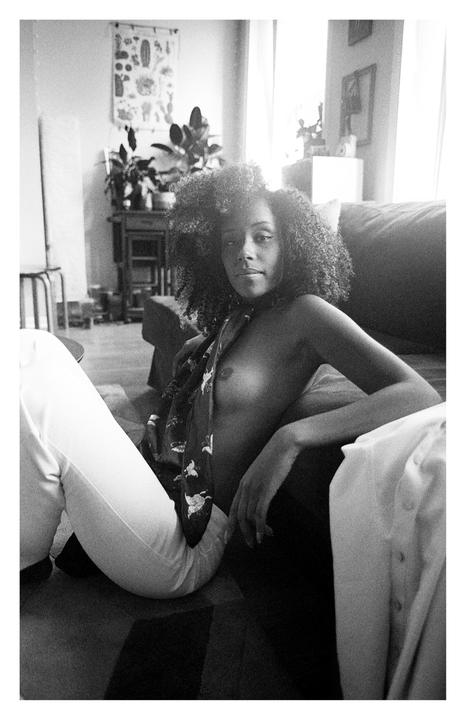 A woman with curly hair sits on the floor in a living room, wearing white pants and a scarf.
