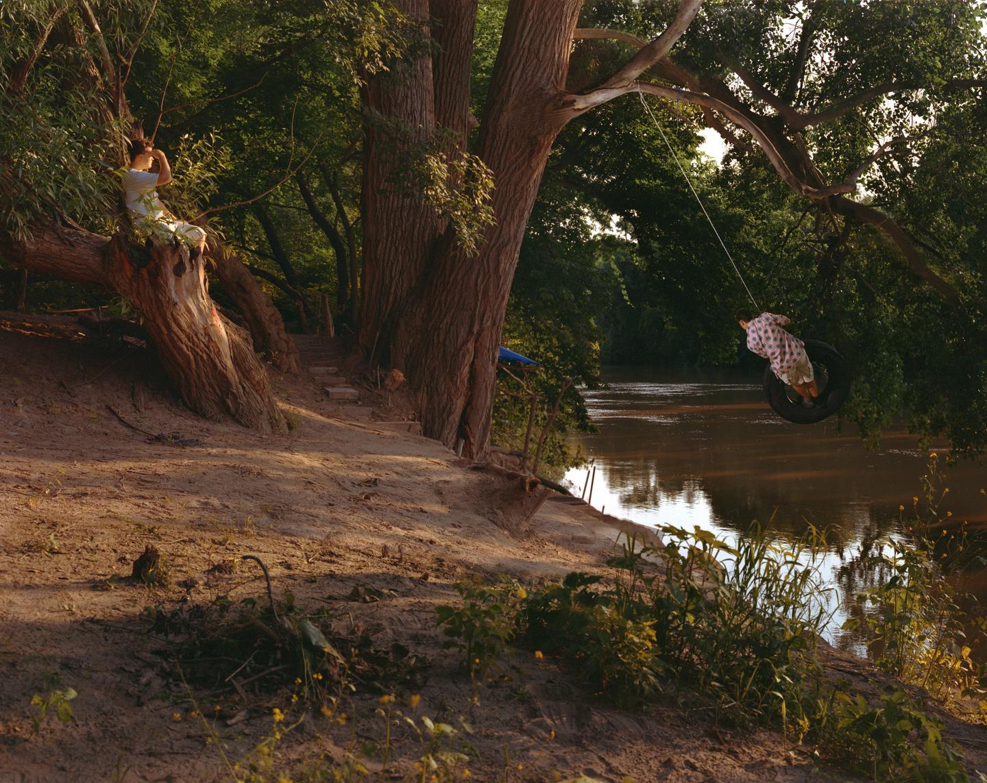A child sits on a tree branch while another child swings on a tire swing over a river.