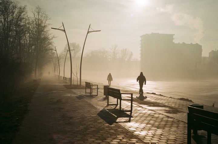 Two people walk along a foggy path lined with benches and streetlights, with a building in the distance.