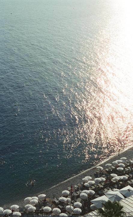 An aerial view shows a beach with many white umbrellas next to the sparkling sea.