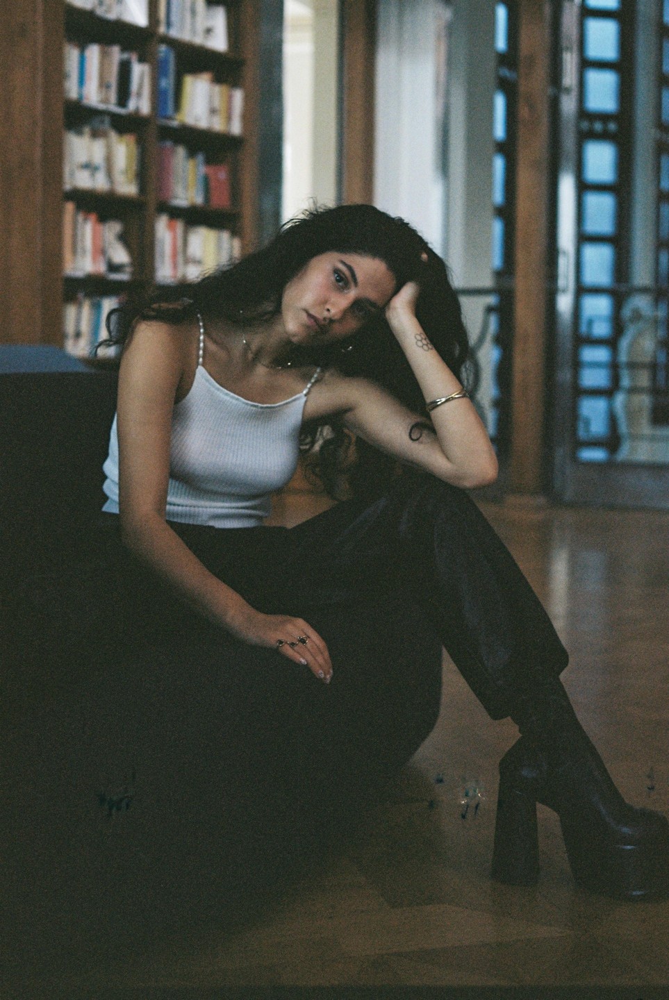 A woman with dark, curly hair sits on the floor in a room with bookshelves in the background.