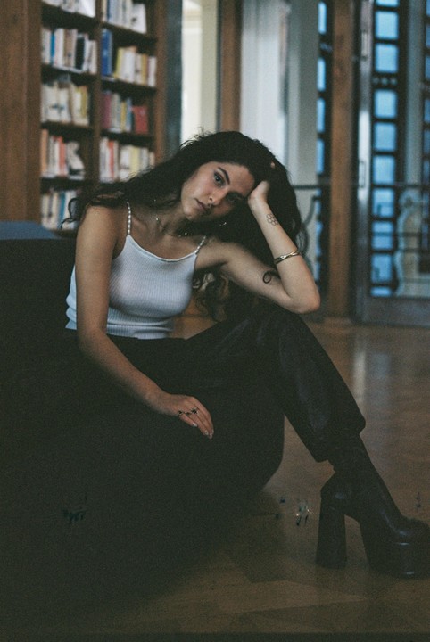 A woman with dark, curly hair sits on the floor in a room with bookshelves in the background.