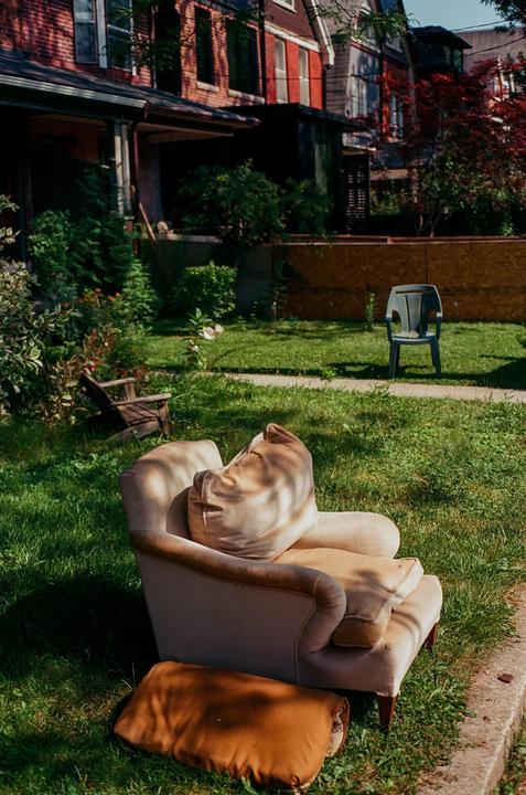 An armchair sits on a grassy lawn in front of a brick house with a plastic chair in the background.