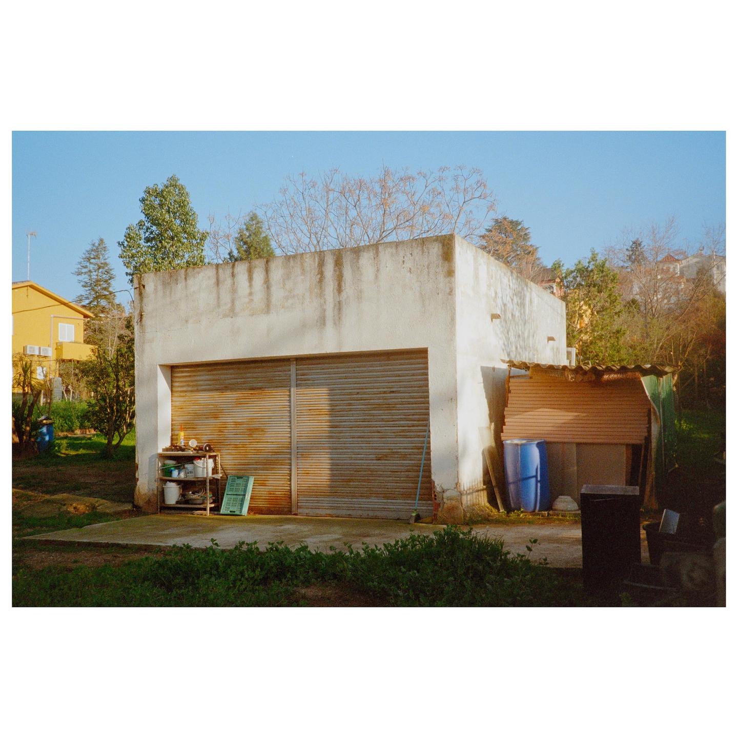 A white garage with a rusty metal door stands in a grassy yard with trees and a yellow building in the background.