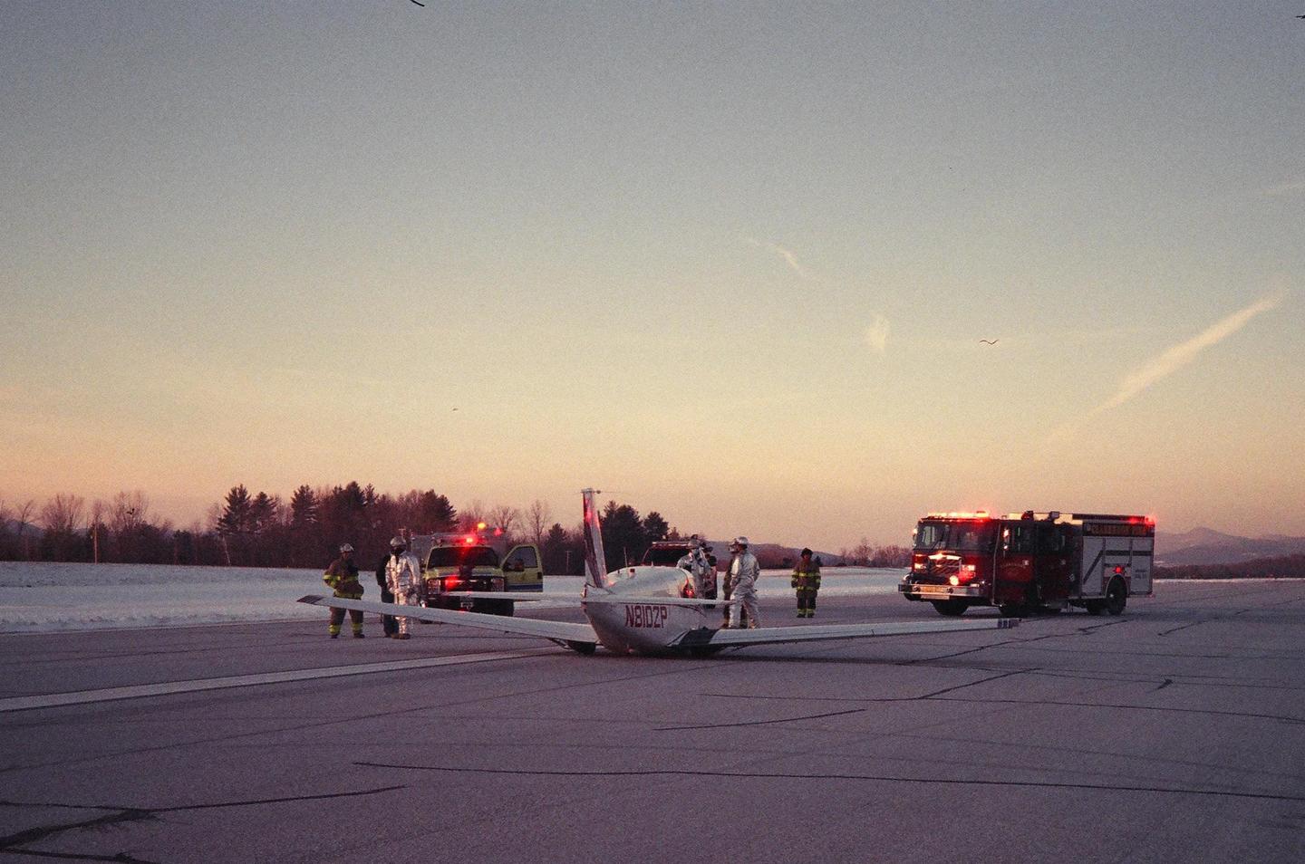 A small plane is on a snowy runway with emergency vehicles and personnel around it at sunset.