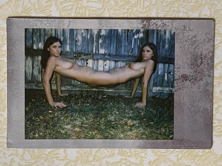 A woman with two heads and four arms is posed on her hands and knees in front of a wooden fence.