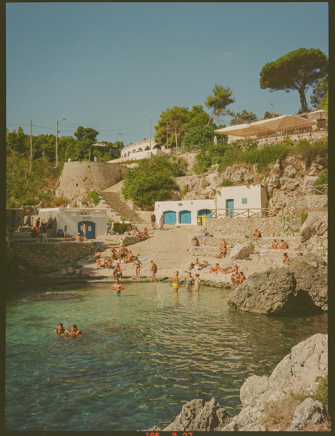 People swim and sunbathe in a rocky cove with white buildings and green trees on the hillside.
