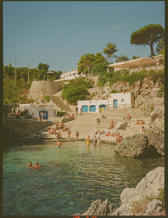 People swim and sunbathe in a rocky cove with white buildings and green trees on the hillside.