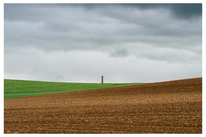 A small tower stands on a green hill under a cloudy sky, with a brown field in the foreground.