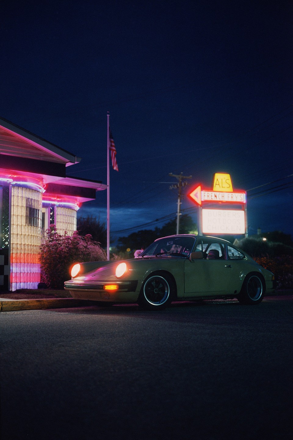A vintage car is parked at night in front of a diner with neon lights and an American flag.