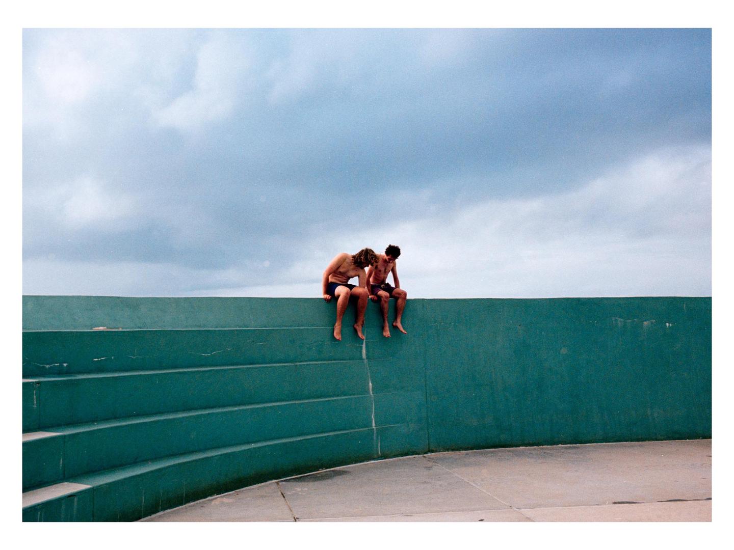 Two men sit on a teal wall under a cloudy sky.