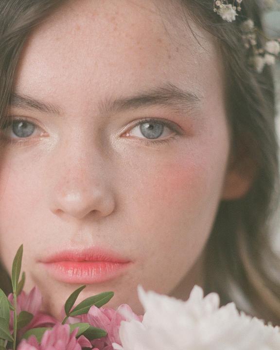 A young woman with flowers in her hair and in the foreground looks directly at the viewer.