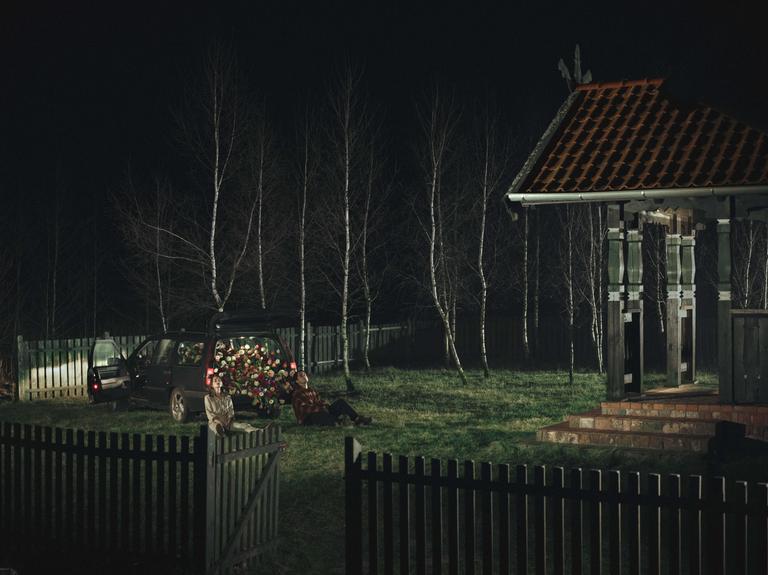 A couple sits on the grass next to a car filled with flowers at night, with a small building and trees in the background.