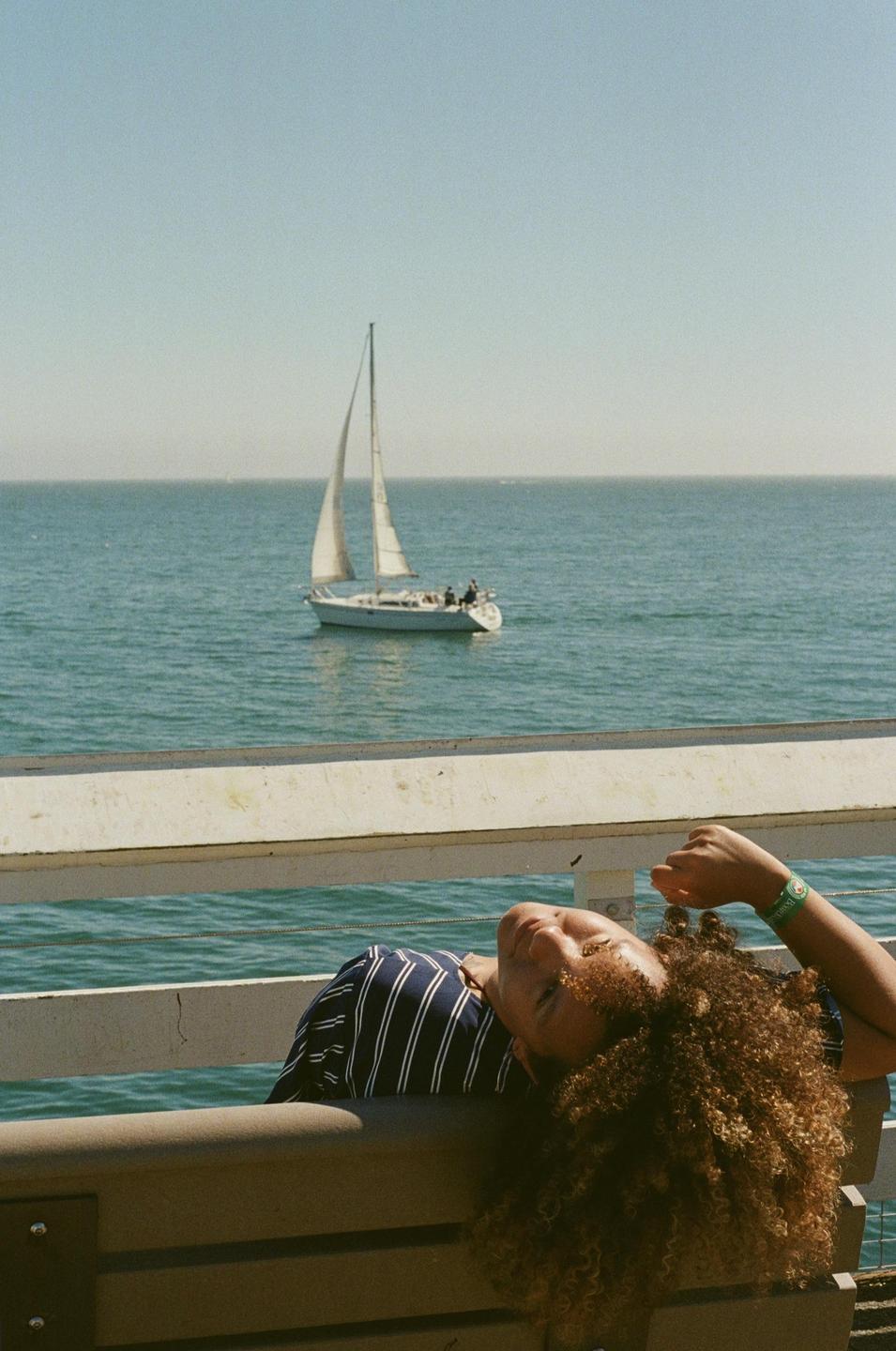 A woman with curly hair is lying on a bench on a pier with a sailboat on the water in the background.