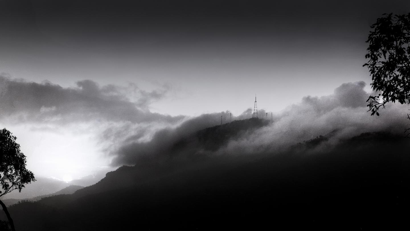 A mountain range is covered in fog with communication towers visible on the peaks and a tree branch in the foreground.