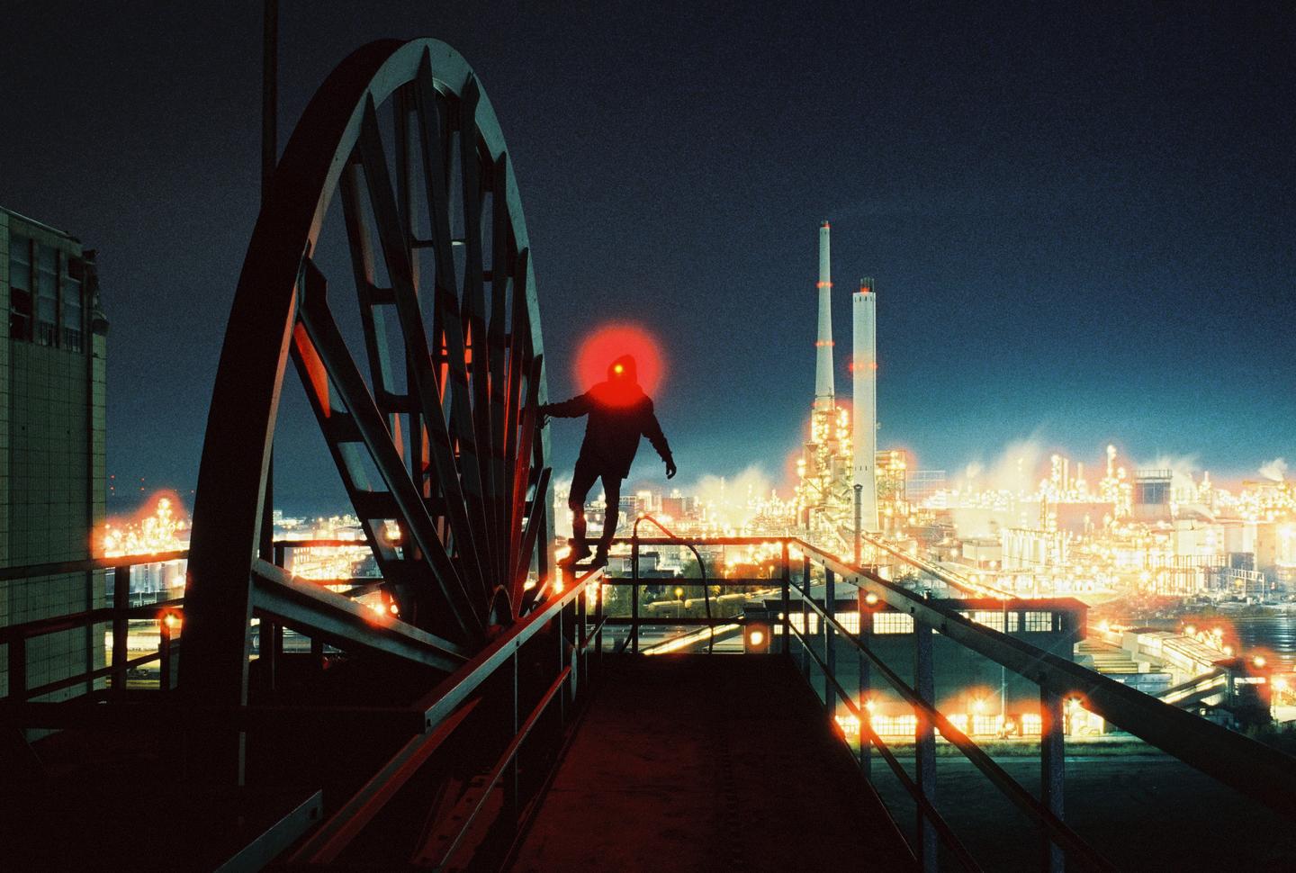 A person stands on a metal structure with a large wheel, overlooking an industrial complex at night.