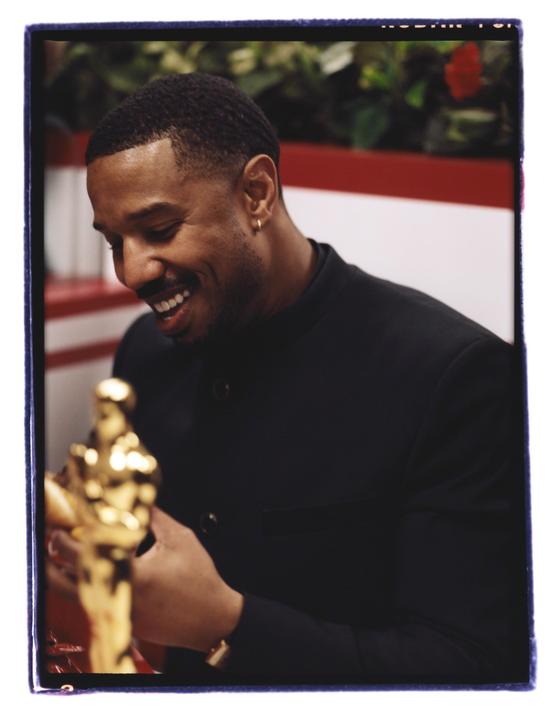 A man in a black jacket smiles while holding a golden statuette.
