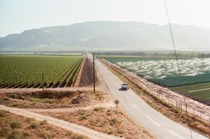 A white pickup truck drives on a road between a vineyard and a field with sprinklers, with mountains in the background.
