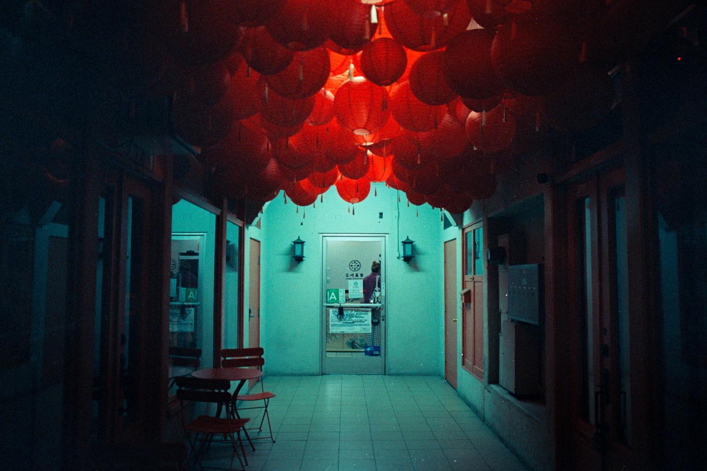 A hallway is decorated with many red lanterns hanging from the ceiling and a person is visible through a glass door at the end of the hall.