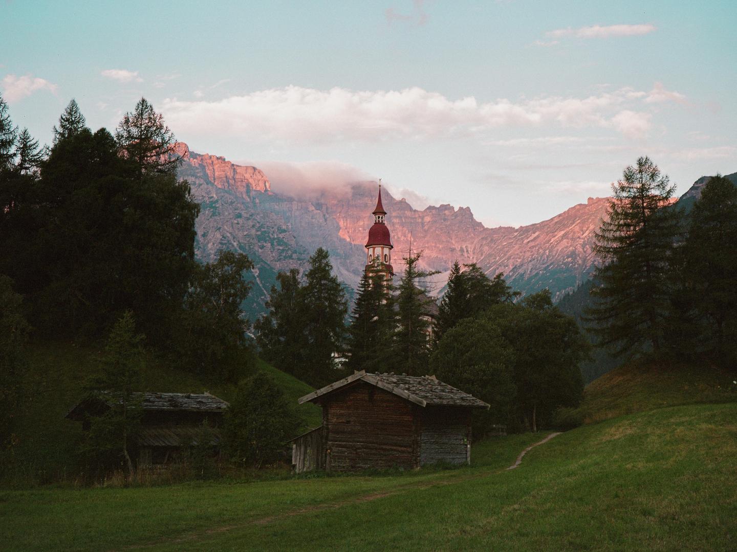 A small wooden cabin sits in a grassy field with a church steeple and mountains in the background.