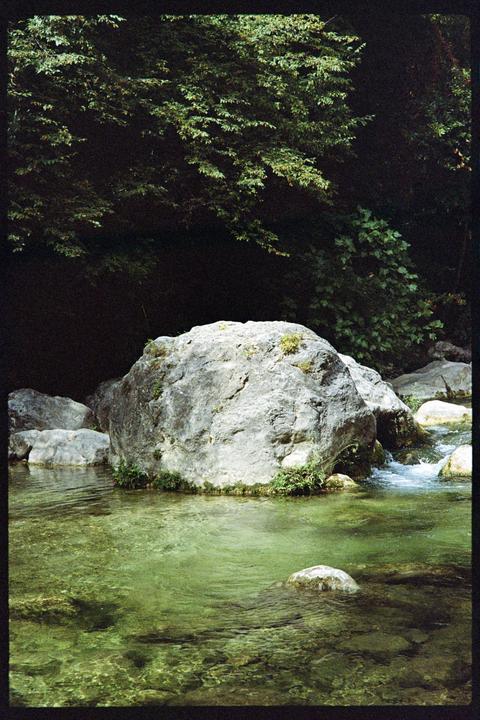 A large rock sits in a clear green river with trees in the background.