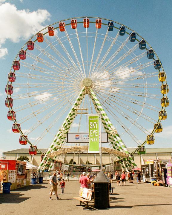 A large Ferris wheel with colorful gondolas stands tall against a blue sky with scattered clouds, overlooking a fairground bustling with people and food stalls.