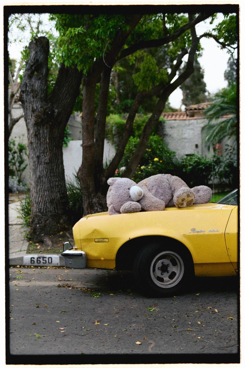 A large teddy bear rests on the hood of a yellow car parked on a street.