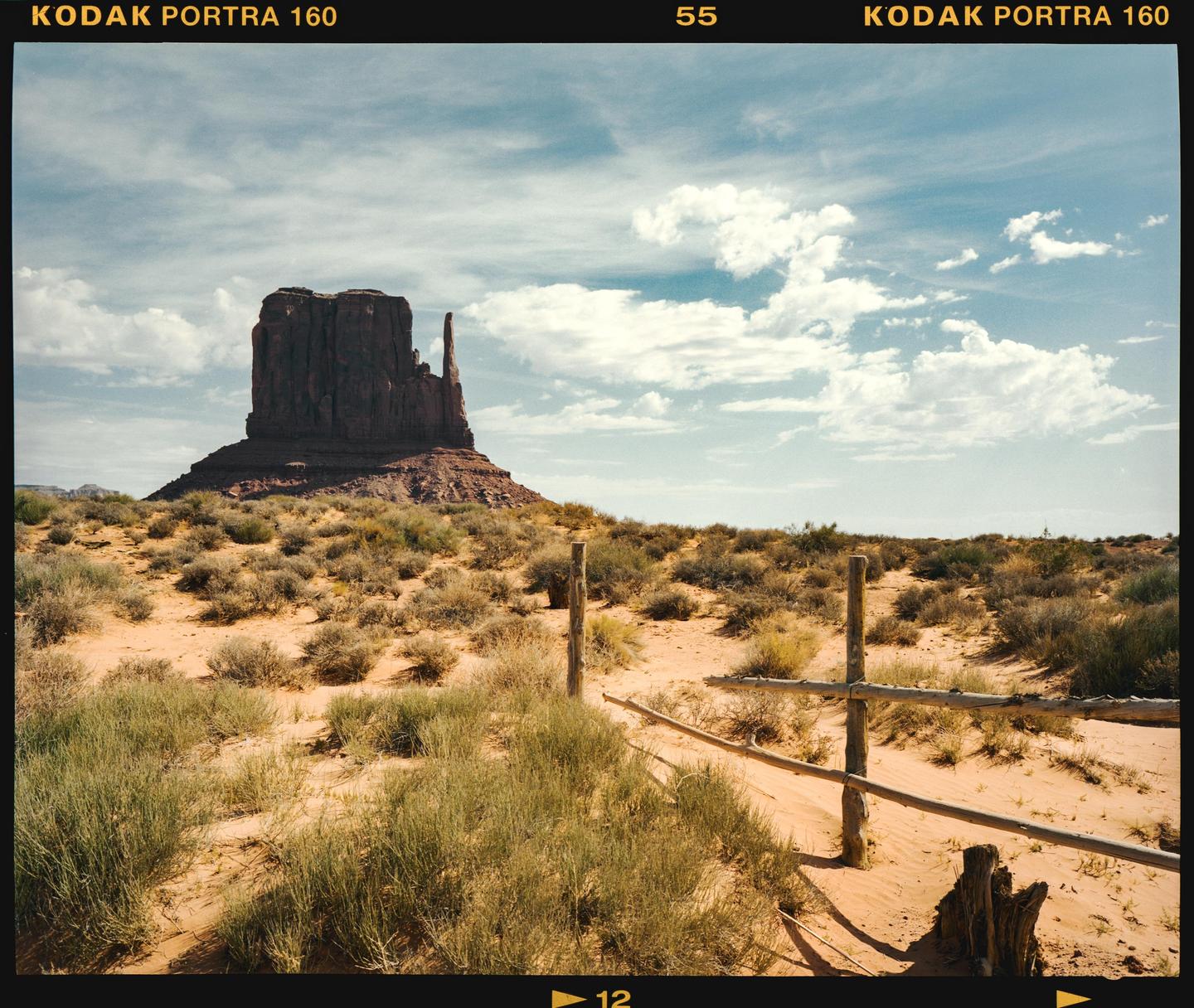 A large rock formation rises from a desert landscape with a wooden fence in the foreground.