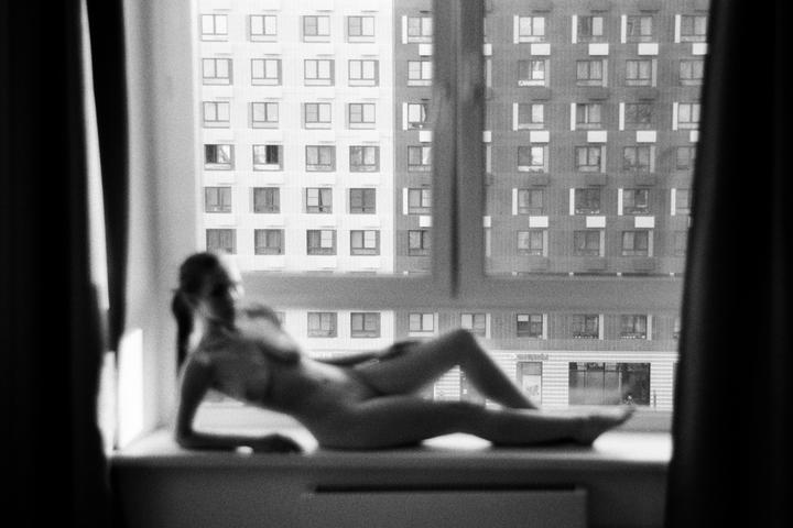 A blurry nude woman reclines on a windowsill with a tall building visible outside the window.