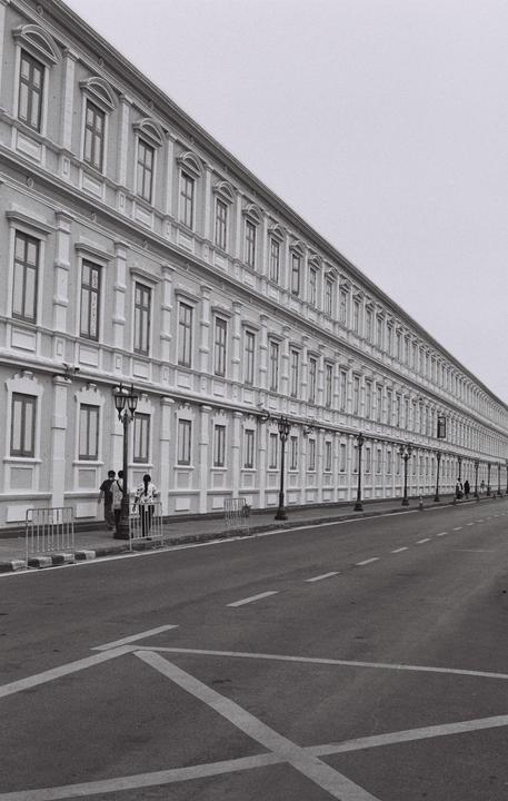 A long, multi-story building with many windows lines a street with a few people walking on the sidewalk.