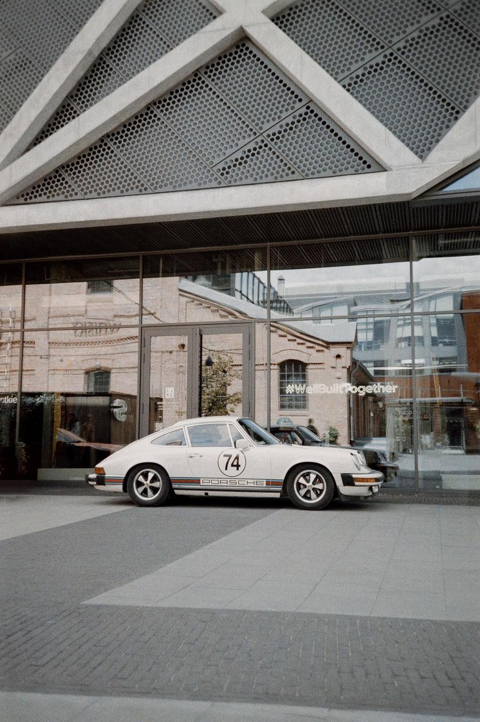A white Porsche with the number 74 on its side is parked in front of a modern building with large glass windows.
