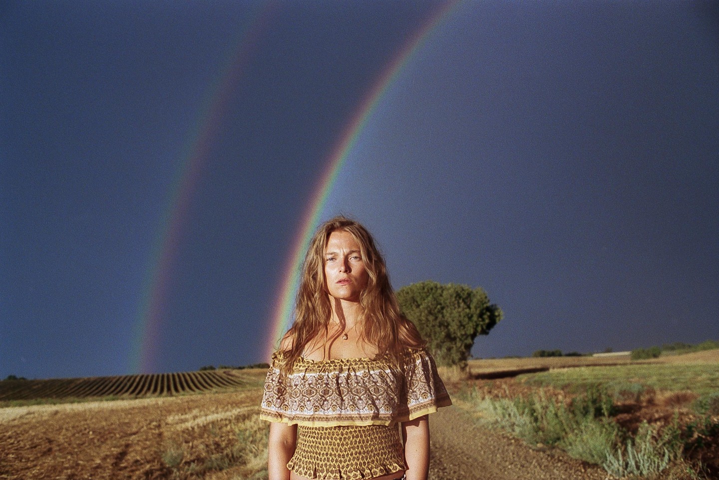 A woman stands in a field under a dark sky with a double rainbow.