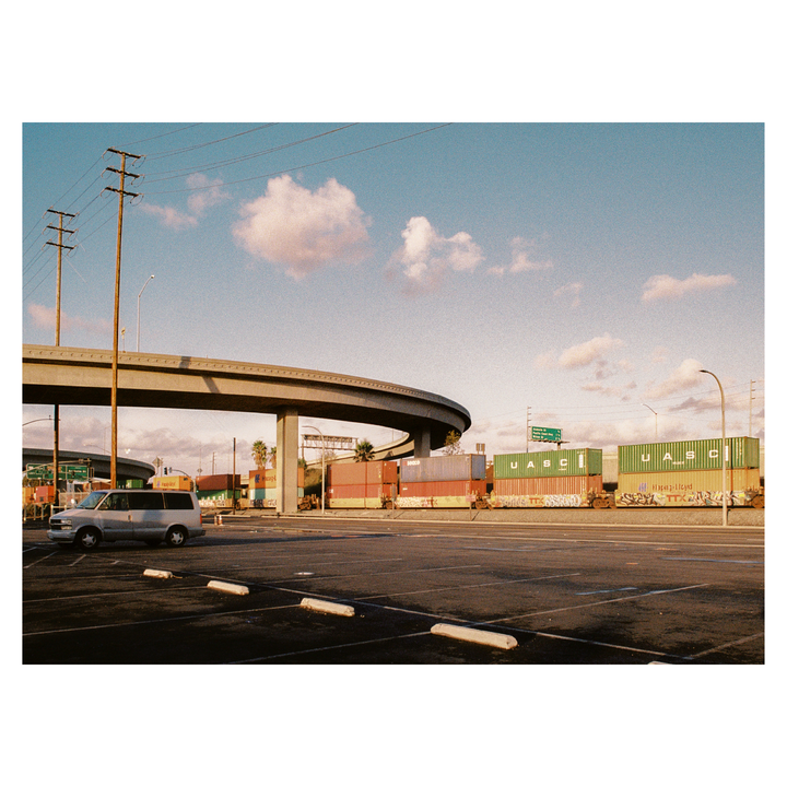 A silver van is parked in an empty lot next to a highway overpass and shipping containers on a train.