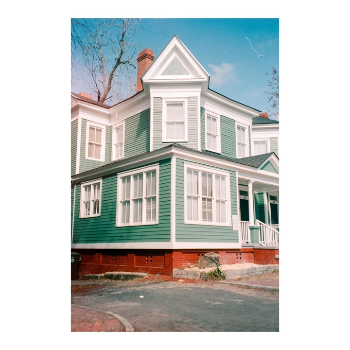 A large green house with white trim and a red brick foundation stands under a clear blue sky.
