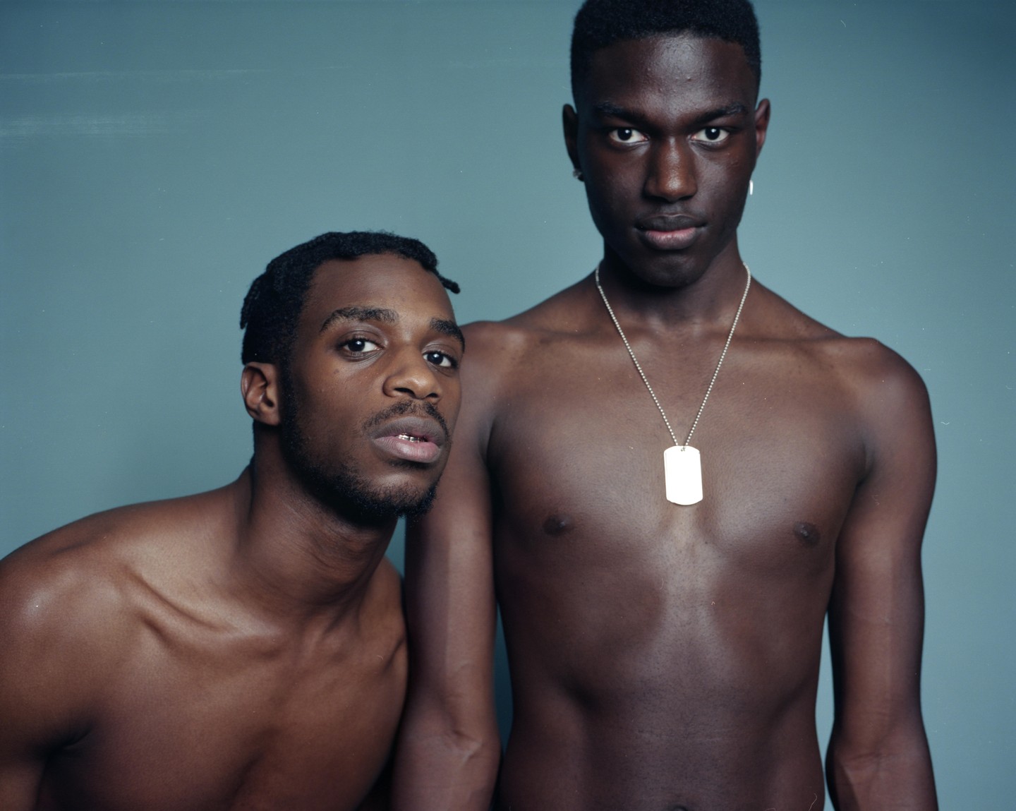 Two men pose shirtless against a blue background, one with a dog tag necklace.