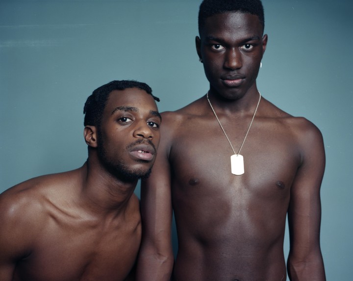 Two men pose shirtless against a blue background, one with a dog tag necklace.