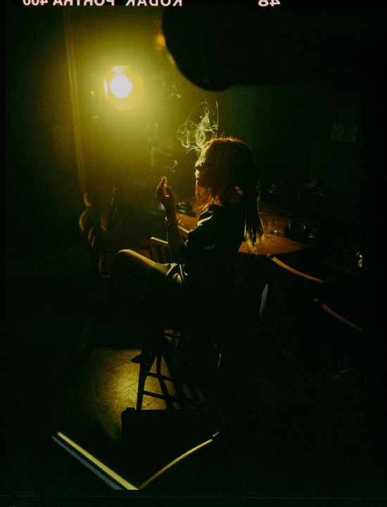 A woman sits in a dark room, smoking a cigarette, with smoke curling around her head.