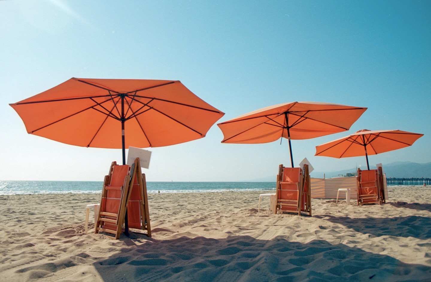Three orange beach umbrellas stand on a sandy beach next to the ocean under a clear sky.