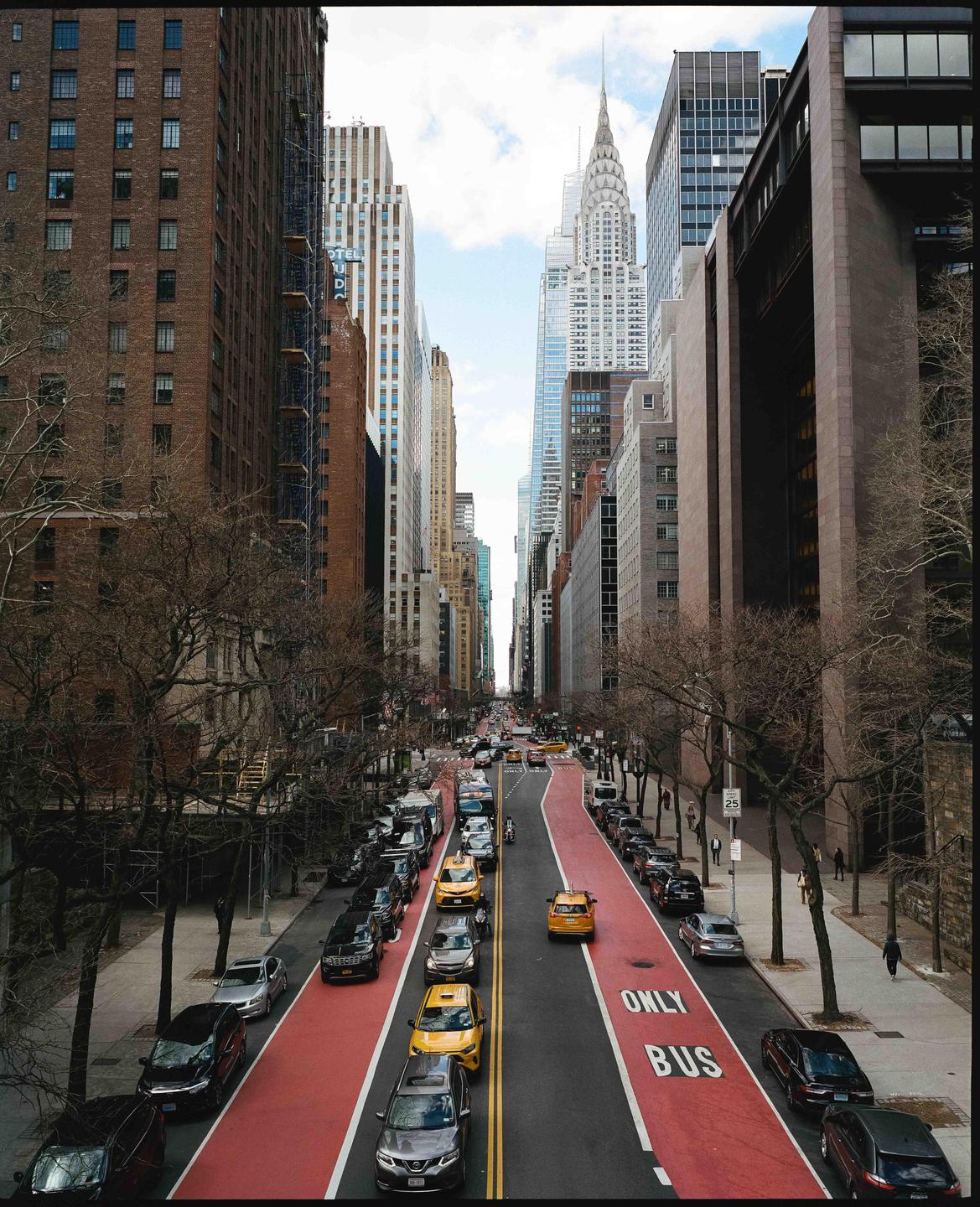 A street in New York City is filled with cars and taxis, with tall buildings and the Chrysler Building in the background.
