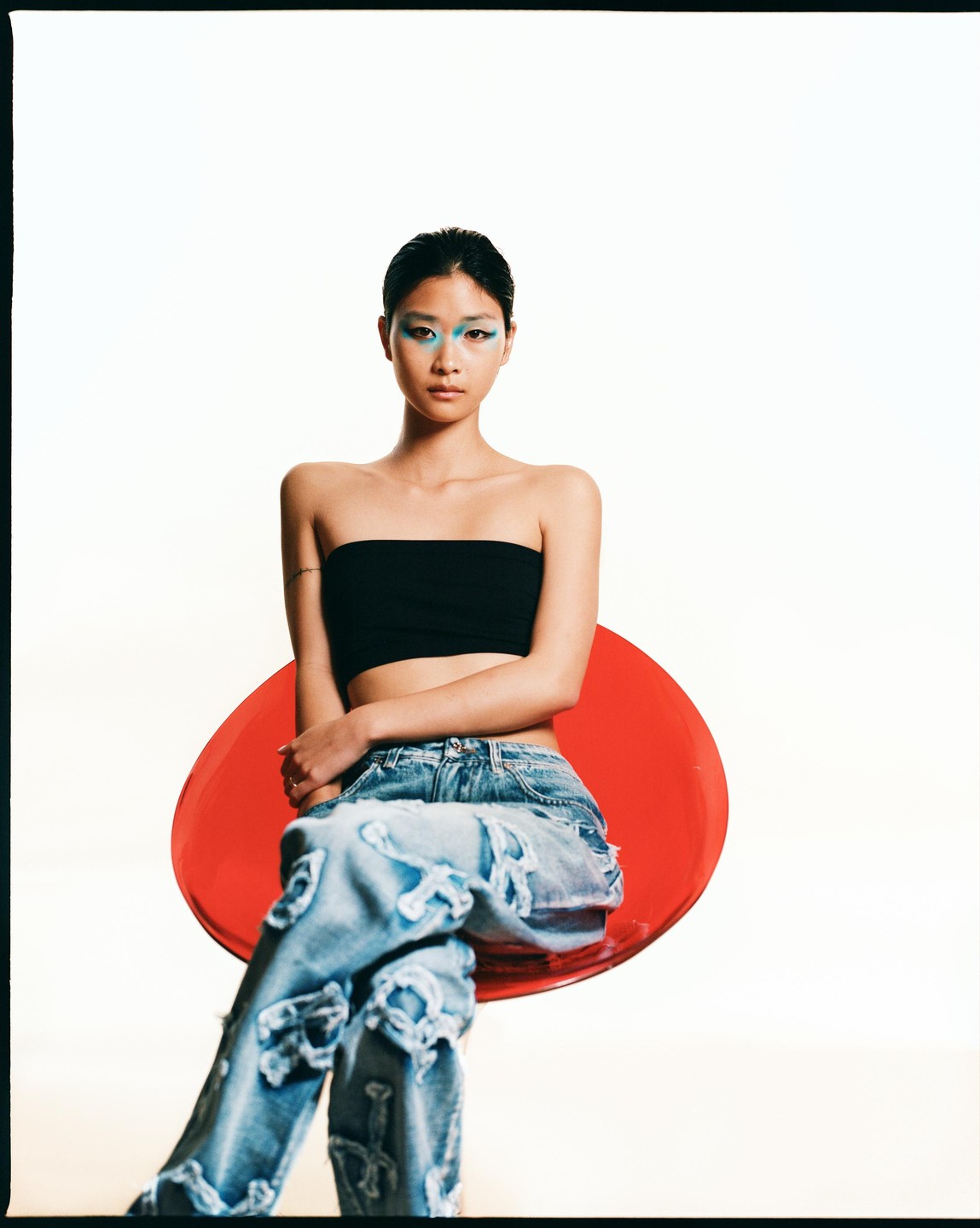 A woman with blue eye makeup and ripped jeans sits on a red chair against a white background.