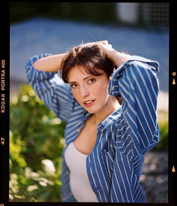 A woman with brown hair and a blue striped shirt poses with her hands behind her head.