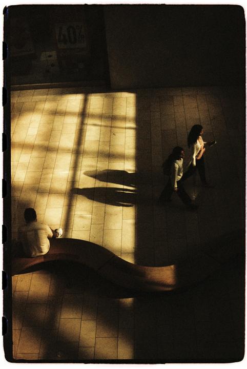 A man sits on a curved bench while two people walk past, all partially illuminated by a window's light.